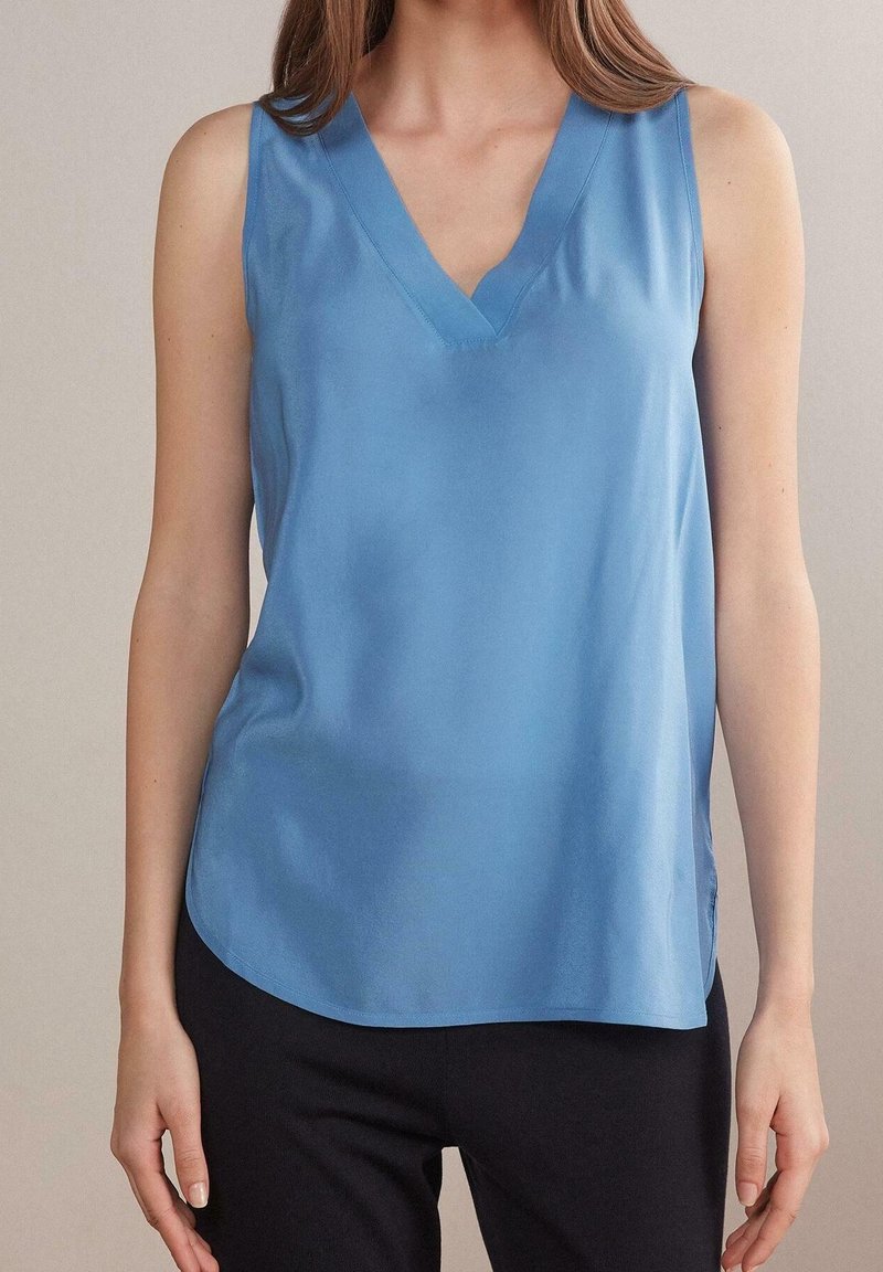 Femme portant un chemisier sans manches bleu clair à col en V et un pantalon noir, avec une peau claire et de longs cheveux bruns visibles.