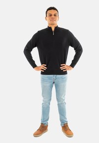 Maglione nero con zip, aderente, polsini a coste, indossato con jeans in denim blu chiaro e stivali marroni. Il modello sta in piedi con le mani sui fianchi.