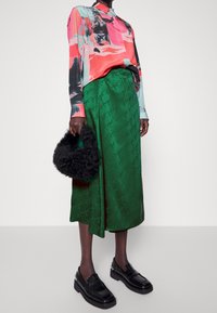 Camisa de manga larga colorida con patrones abstractos, combinada con una falda de satén verde con un sutil estampado de logo, accessorios con un bolso de piel de pelo negro.