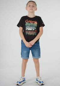 T-shirt noir avec un graphique "California Holiday", short en denim bleu avec des ourlets roulés, et baskets blanches avec des accents bleus. Design simple.