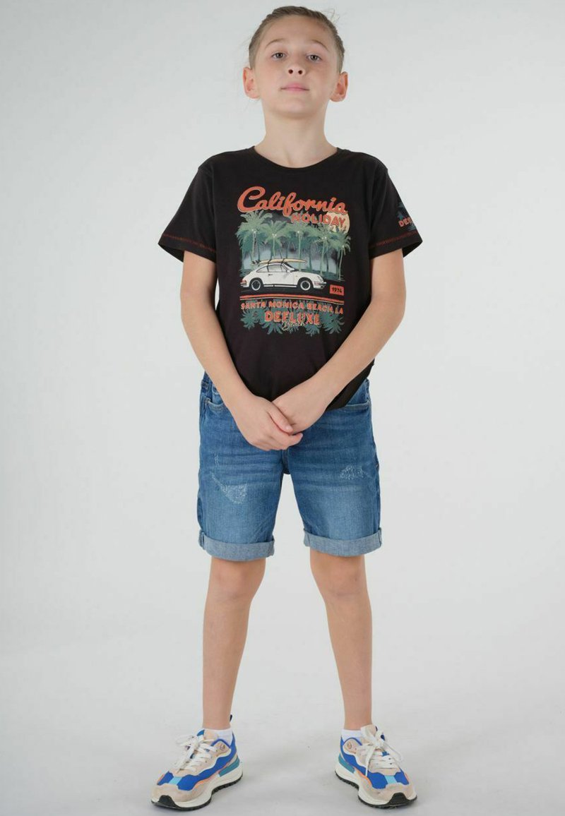 T-shirt noir avec un graphique "California Holiday", short en denim bleu avec des ourlets roulés, et baskets blanches avec des accents bleus. Design simple.