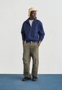 Φούτερ με φερμουάρ σε ναυτικό μπλε χρώμα, χακί παντελόνι cargo με πλαϊνές τσέπες, μαύρα παπούτσια και ανοιχτό μπεζ καπέλο. Απλός, λειτουργικός σχεδιασμός.