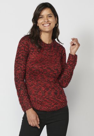 Pull en tricot rouge et noir avec un col rond, des manches longues et un motif texturé. La coupe est ajustée, mettant en valeur la taille.