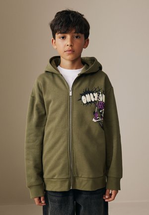 Niño con el pelo corto y oscuro que lleva una sudadera con capucha verde oliva con cremallera y estampado gráfico y vaqueros oscuros, de pie frente a un fondo liso beige.
