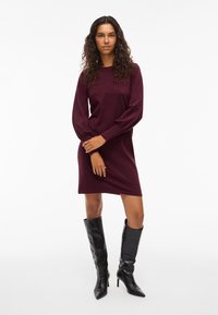 Burgundy mekko, jossa on pitkät, puffatut hihat ja pyöreä kaulus, yhdistettynä polveen ulottuviin mustiin korkokenkiin. Sileä kangas.