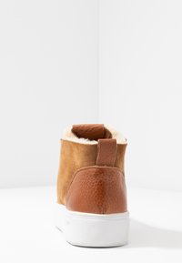 Brown suede and leather ankle boot with faux fur lining, white rubber sole, and a rear pull tab for easy wear. Smooth and textured finish.