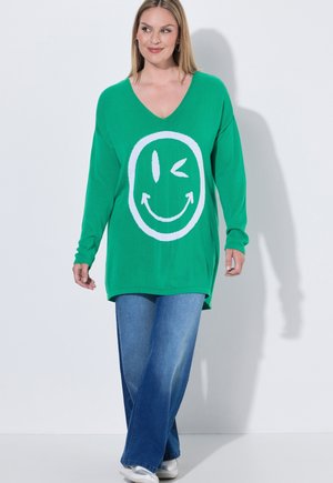 Kvinde iført en grøn langærmet sweater med et hvidt blinkende smiley, blå jeans og hvide sneakers går fremad.