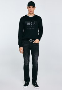 Mann steht und trägt eine schwarze Mütze, einen schwarzen Pullover mit dem Schriftzug "BIG STAR", schwarze Jeans, einen schwarzen Gürtel und schwarze Turnschuhe mit weißen Sohlen.