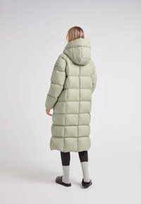 Cappotto puffer lungo verde chiaro con cappuccio, caratterizzato da un design trapuntato, ampi pannelli e lunghezza fino al polpaccio, abbinato a leggins scuri e calze.
