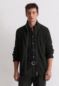 Donkergroene ritssluiting cardigan met ribbelstructuur, hoge kraag en opgerolde manchetten. Gekoeld over een zwart shirt, met een zilveren rits en accenten.