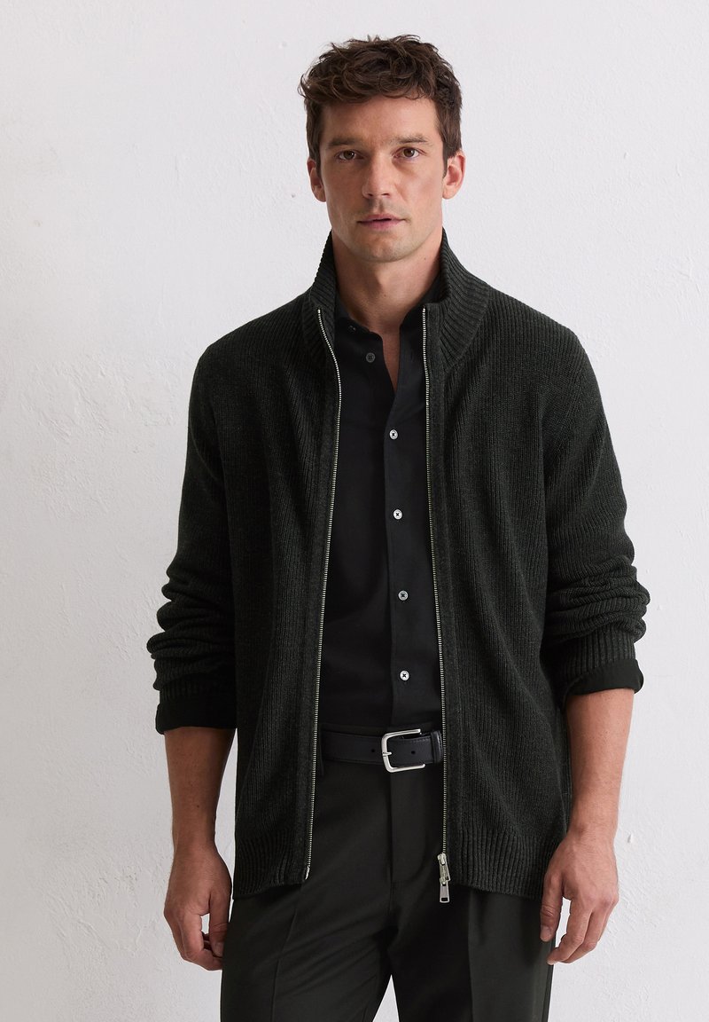 Donkergroene ritssluiting cardigan met ribbelstructuur, hoge kraag en opgerolde manchetten. Gekoeld over een zwart shirt, met een zilveren rits en accenten.