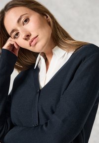 Mörkblå stickad cardigan med V-ringning och knappstängning, bärs över en vit skjorta med krage. Har ribbade muddarna och en texturerad tyg.