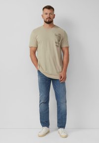 Beige katoenen t-shirt met een ronde hals en korte mouwen, met een grafische print op de linkerkant van de zak. Gecombineerd met lichtblauwe jeans en witte sneakers.