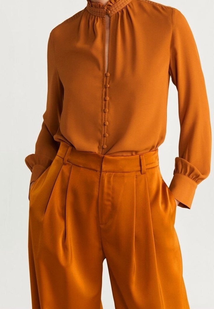 Blouse orange brûlé avec détails à boutons et col montant ; associée à un pantalon plissé assorti, tous deux en tissu léger et lisse.