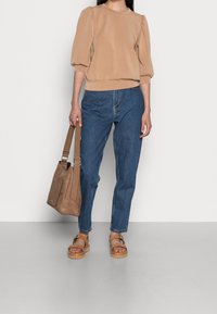 Sudadera beige de manga abullonada, vaqueros de tela vaquera azul y sandalias de cuero color tostado. Lleva un bolso marrón texturizado con una correa.