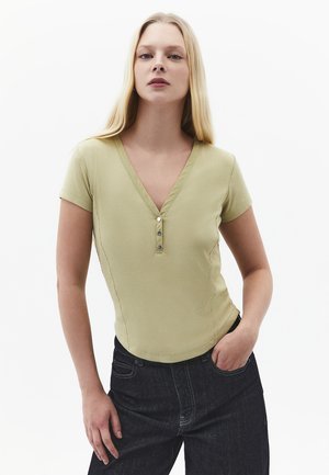 Jonge vrouw met lang blond haar, gekleed in een lichtgroen shirt met korte mouwen, V-hals en knopen, en een donkere spijkerbroek, poserend met één hand in haar zak.