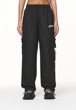 Femme portant un pantalon cargo noir à jambes larges avec le texte "PEGADOR" et des baskets noires, debout devant un fond blanc.