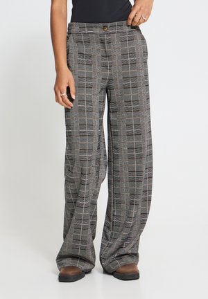 Femme portant un pantalon large à carreaux gris avec fermeture à boutons et des chaussures marron, debout devant un fond clair uni.