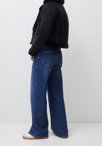 Personne portant une veste en peau retournée noire, un jean large bleu et des baskets blanches, debout devant un fond blanc uni.