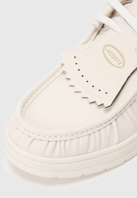 Detailní záběr na béžovou koženou obuv Lacoste s vyšívanými detaily, dekorační perforovanou klapkou a bílou texturovanou podrážkou.