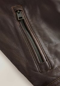 Braune Lederjacke mit einer strukturierten Oberfläche, ausgestattet mit einem Metallreißverschluss entlang der Naht, der Handwerkskunst und Langlebigkeit hervorhebt.