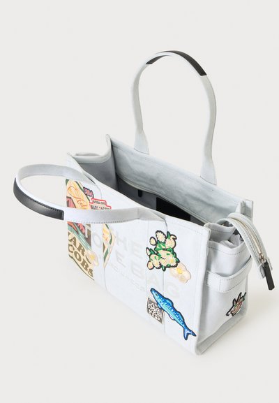 Sac cabas blanc avec poignées noires, décoré de patchs colorés dont des fleurs, un poisson et divers logos ; poche zippée visible.