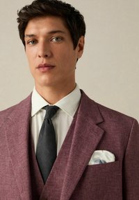 Bordeauxfarbener Blazer mit strukturiertem Finish, weißes Hemd und schwarze Krawatte. In der Brusttasche befindet sich ein gemusterter Einstecktuch.