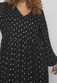 Vestido negro con estampado de hojas plateadas, escote en V, falda plisada y mangas largas transparentes. Textura suave con un diseño fluido.