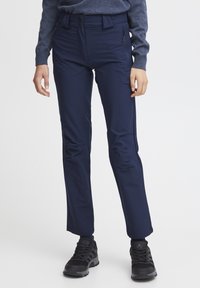 Pantalon bleu marine à la texture lisse, avec une taille haute, deux boutons, des poches zippées sur les côtés et des jambes fuselées, associé à des chaussures noires.
