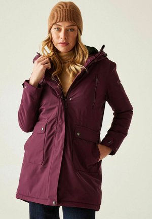 Kobieta w bordowej parce z wieloma kieszeniami i beżowej dzianinowej czapce, poprawiająca kołnierz kurtki, stojąca na tle jednolitego tła.