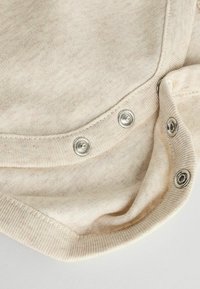 Ljust beige tyg med en mjuk textur, med silverfärgade tryckknappar längs kanten och räfflad trim.