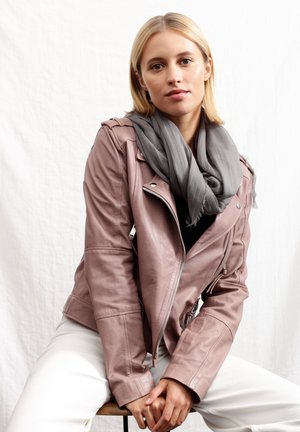 Veste de biker en cuir rose avec des détails de fermeture éclair en argent, associée à une écharpe grise. Le modèle est assis sur un tabouret devant un fond neutre.