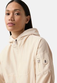 Veste beige clair avec capuche, dotée d'une fermeture éclair, d'une poche avant et d'un patch logo sur la manche. Tissu lisse et léger.