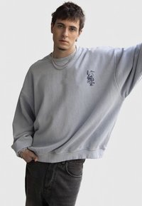 Lyseblå oversized sweatshirt med nedfalne skuldre, ribbede mansjetter og et svart trykt design på brystet. Kombinert med mørke jeans.