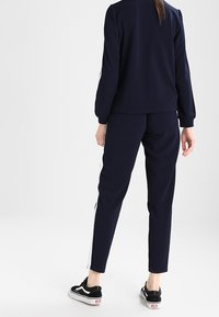 Navy tracksuit dengan lengan panjang dan pergelangan tangan elastis. Celana tapered memiliki garis samping putih. Dipadukan dengan sepatu sneaker hitam.