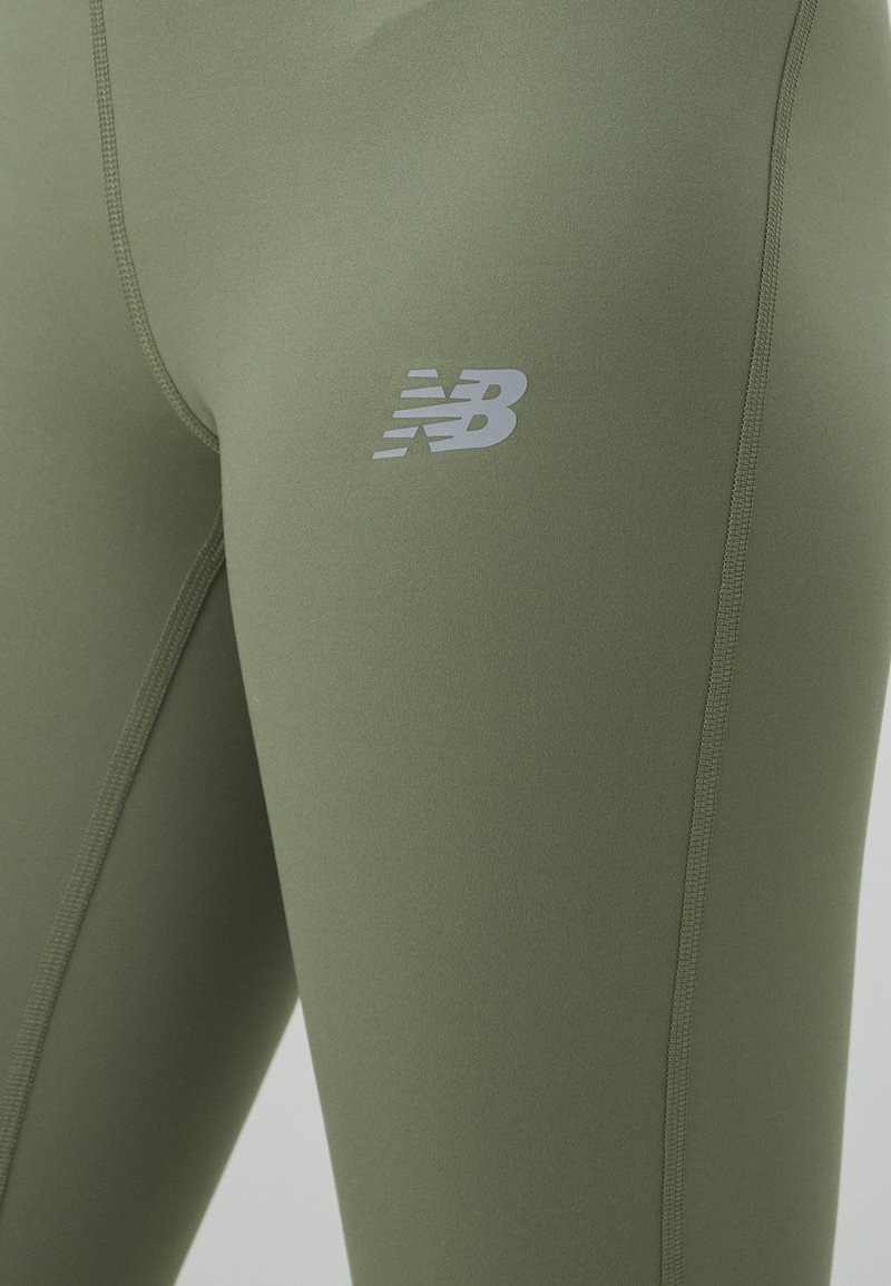 Leggings de color verde oliva hechos de una tela elástica. Presentan un logo plateado de New Balance en el costado y detalles de costura visibles.