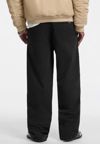 Black wide-leg pants made from durable fabric, featuring a smooth texture and two back pockets, worn with a beige bomber jacket.