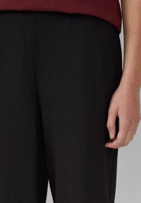 Pantalons noirs texturés avec un design côtelé. Le tissu est léger, avec une coupe ample, et est complété par un haut bordeaux partiellement visible au-dessus.
