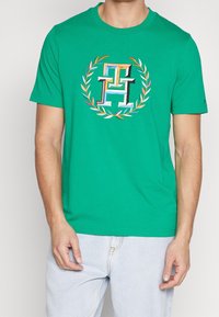Camiseta de algodón verde con un logo bordado colorido en el centro, rodeado por un patrón de corona de laurel. Mangas cortas, cuello redondo.