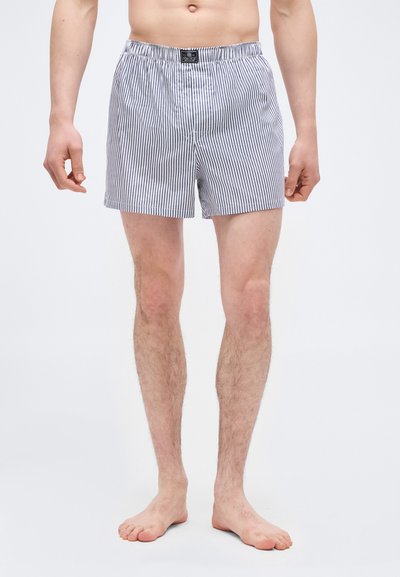 Άνδρας που φορά γκρι και λευκά κατακόρυφα ριγέ μποξέρ, όρθιος ξυπόλητος μπροστά σε απλό λευκό φόντο.