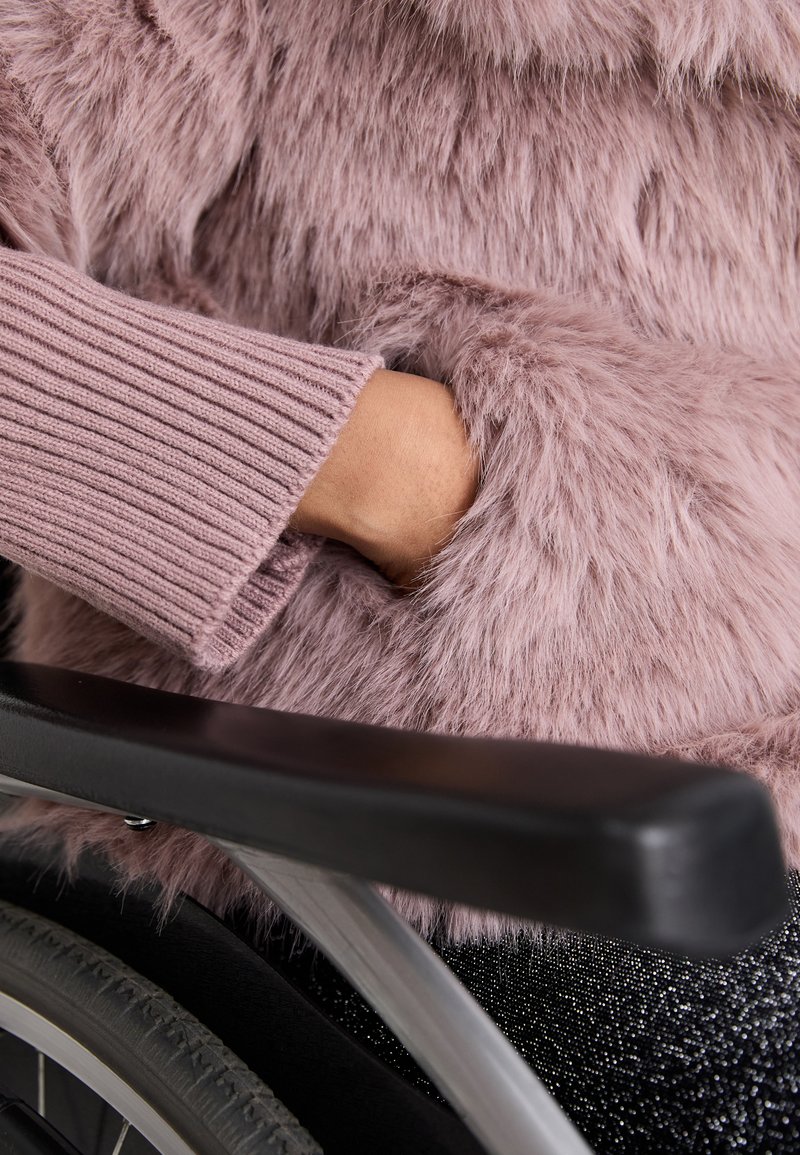 Giacca in pelliccia sintetica di morbido rosa con maniche a costine. Una mano è infilata nella tasca accanto al bracciolo di una sedia a rotelle. Texturizzata e accogliente.
