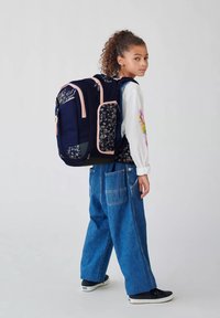 Zaino navy con motivo floreale e dettagli rosa, dotato di diverse tasche e schienale imbottito, indossato sopra una camicia bianca e jeans blu.