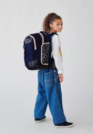 Sac à dos marine avec motif floral et accents rose, doté de plusieurs poches et d'un dos rembourré, porté sur une chemise blanche et un jean bleu.