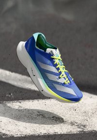 Chaussure de course bleue avec tige en mesh, accents verts et lacets jaunes vifs. Présente une finition texturée et trois rayures blanches sur le côté.