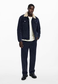 Mornarsko plava traper jakna s kremastim umjetnim krznom na ovratniku, u kombinaciji s odgovarajućim mornarsko plavim hlačama širokih nogavica i crnim cipelama.