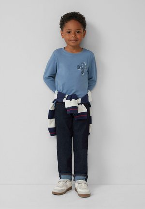 Blaues Langarmhemd mit besticktem Muster, marineblau-weißer gestreifter Pullover um die Taille gebunden, dunkle Jeans und weiße Sneakers.