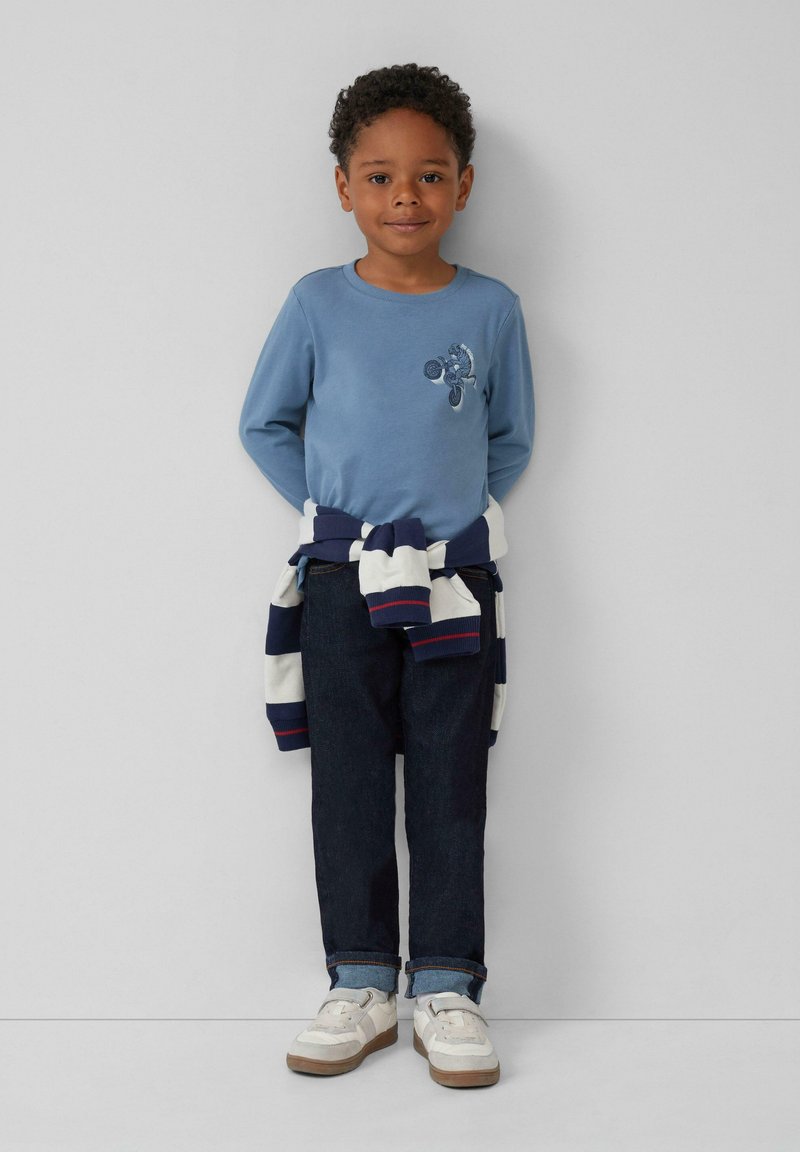 Blauwe langemouw shirt met geborduurd ontwerp, marineblauw en wit gestreepte trui om de taille geknoopt, donkere jeans, en witte sneakers.