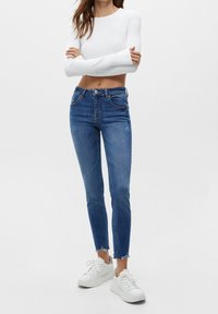 Jeans de mezclilla azul con un corte ajustado, dobladillo deshilachado y diseño clásico de cinco bolsillos, combinados con una blusa blanca de manga larga y zapatillas blancas.