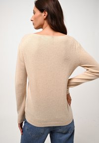 Beżowy, miękki sweter z dzianiny z szerokim dekoltem i długimi rękawami. Posiada luźny krój i ribbowe wykończenie, zestawiony z niebieskimi dżinsami.