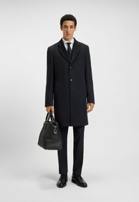 Cappotto navy con un design su misura, due bottoni e tasche laterali, abbinato a una borsa in pelle nera e scarpe nere, indossato sopra un completo.